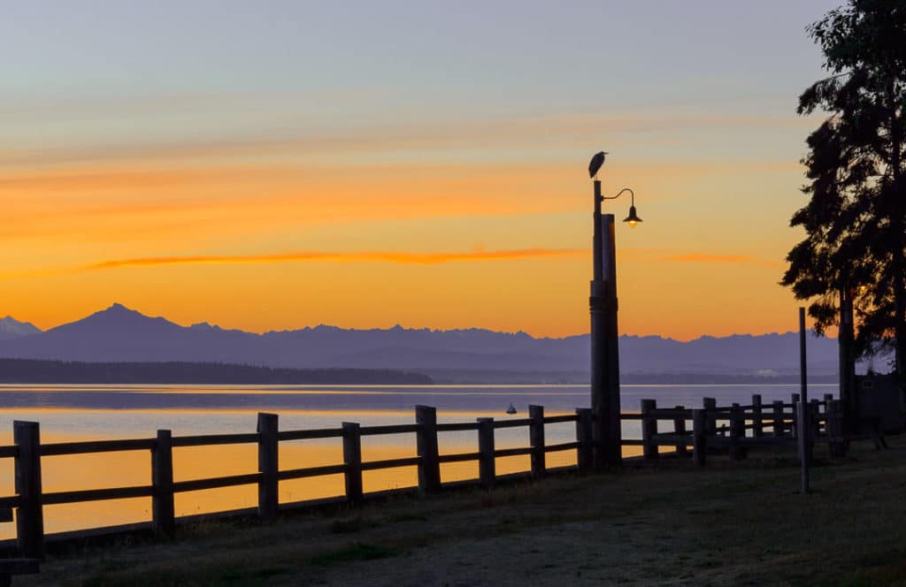 A fence and post are in the foreground with a heron on the post. There are water and mountains behind that and a sunrise behind that.