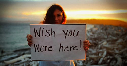 Young woman at sunset on a beach holding a sign that says "Wish you were here!"