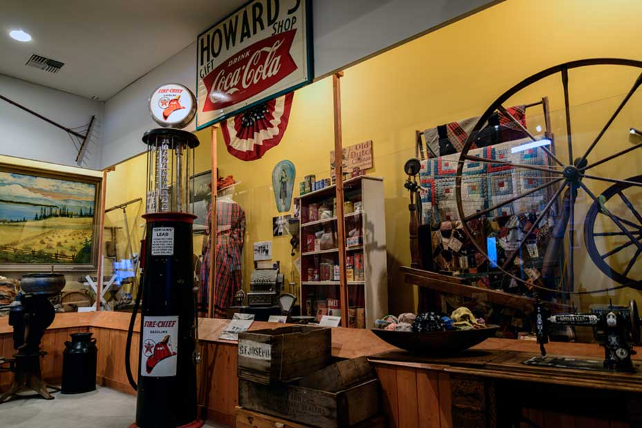 A display case filled with such things as quilts, a cash register, and things you might find in an 19i20's era market. A gas pump sits in front of the display.