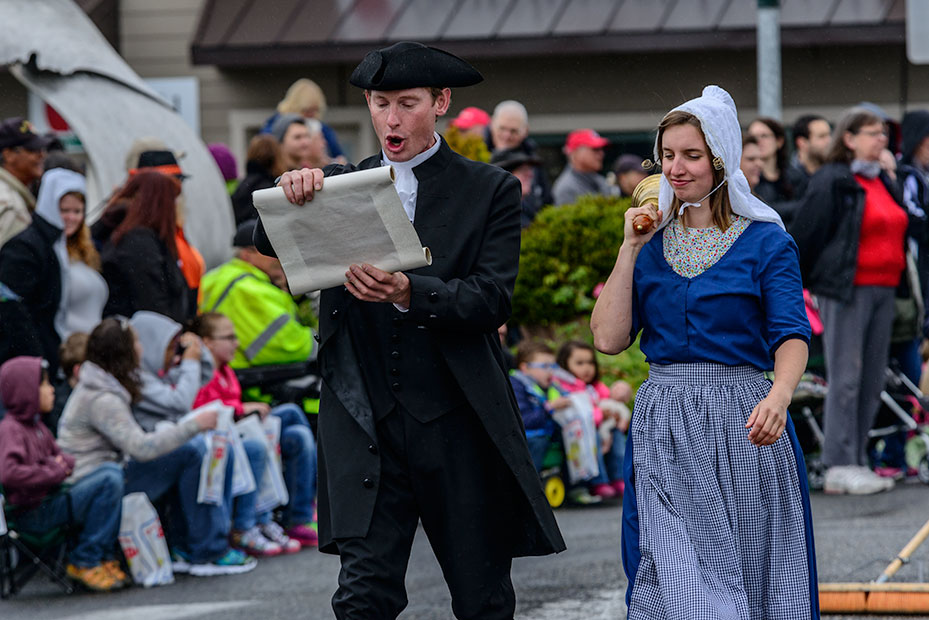 A man and woman in traditional Dutch clothing walk in the parade. He's proclaiming the holiday while reading from a scroll. She's ringing a bell.