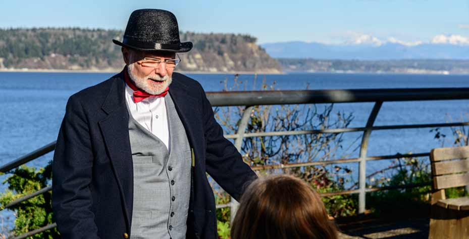 A man in in a dark suit, red bow tie and dress hat answers questions from amateur detectives.