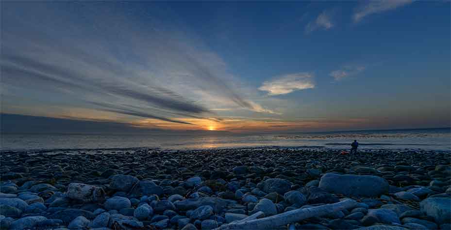 A low-angle view of a rocky beach with a hazy sunset in the background.