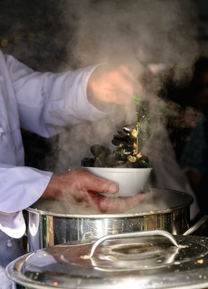 A left hand ladling mussels into a bowl held by the right hand. There is steam all around.