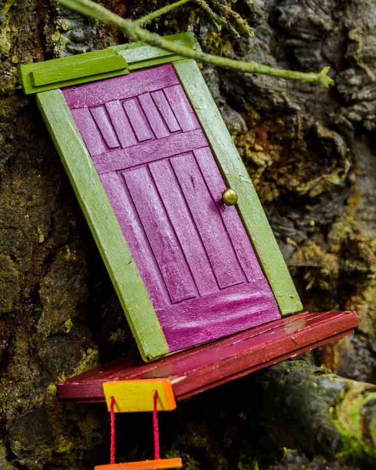 A tiny purple door is surrounded by an avocado green doorframe and sitting on a red porch, sits on a branch and leans against a trunk. A tiny rope latter hangs to one side.