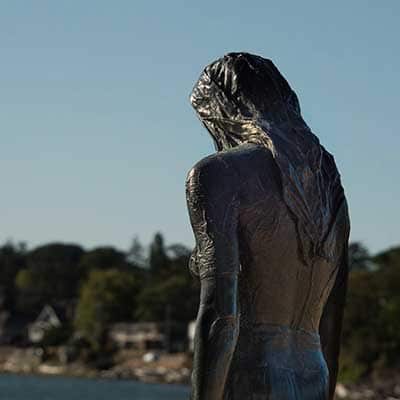 Statue of a woman standing at the shore as seen from behind.