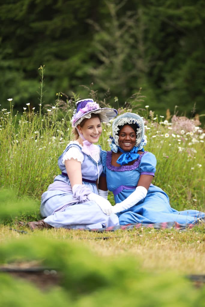 Two women dressed in earth 20th century English dresses sit in the grass and smile. 
