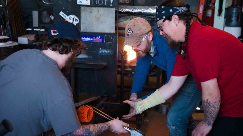 A glass artist and an assistant create a bowl while another man looks on.