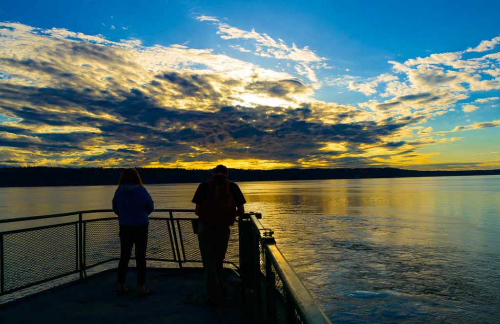 Two people on the deck of a ferry watch the sun set.