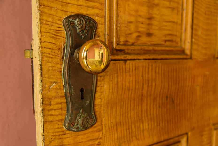 An antique doorknob on a wooden door. The knob, itself, is shiny and reflects the room.