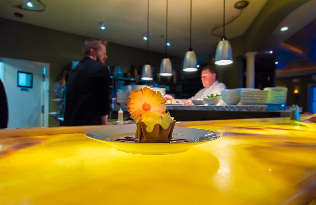 A frilly dessert sits on a counter with two chefs talking to each other in the background.