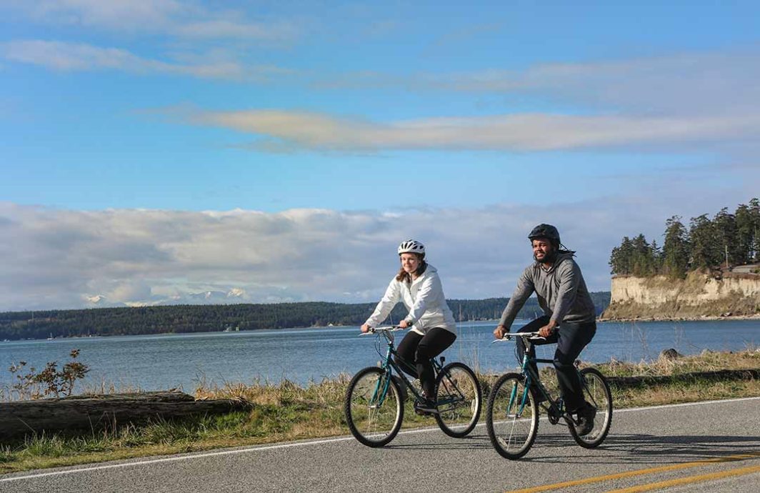 Two people riding bicycles with water and a bluff in the background.