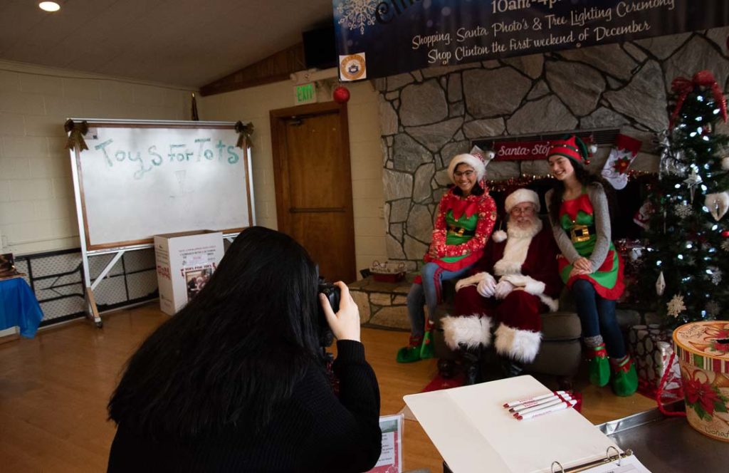 Photographer takes a picture of Santa Claus sitting on a hearth with two elves on either side.