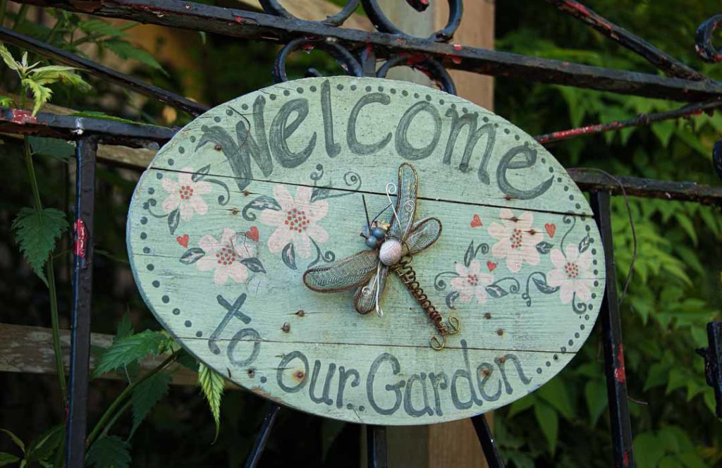 Wooden sign says Welcome to our garden. There is dragonfly made out of metal on it.
