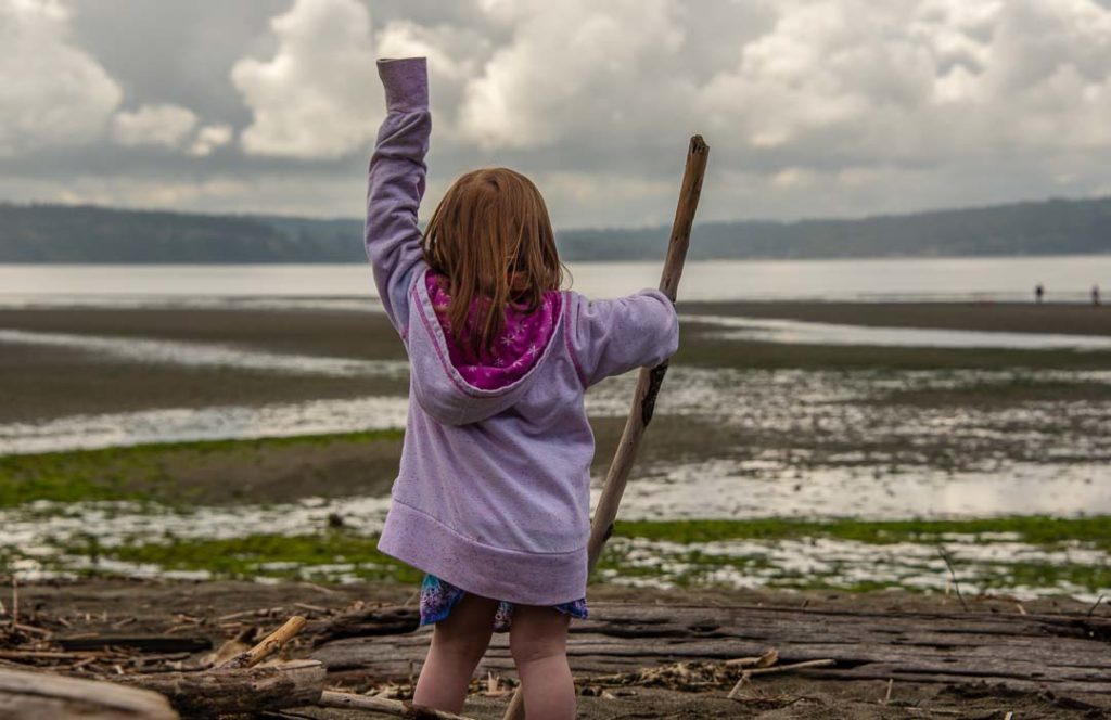 Young girl in a purple jacket raises her left hand towards the sea and hold a stick in her right.