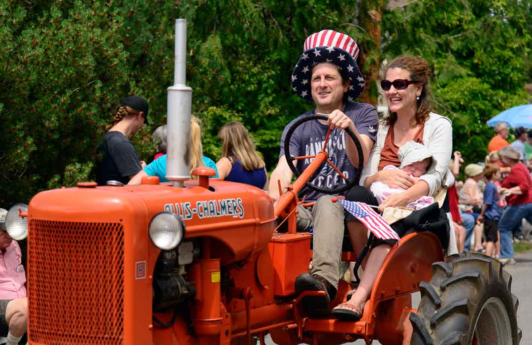 Young couple with sleeping baby driving an old tractor in a parade.
