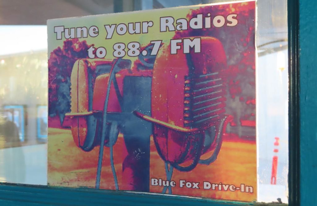 Sign says "Tune your radios to 88.7 FM" and shows old fashioned drive in speakers.