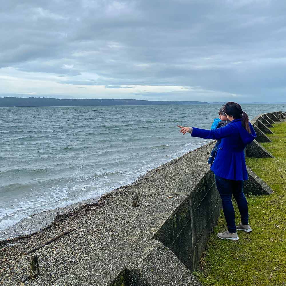 Woman in a blue coat holds an infant next to a seawall on a blustery day.