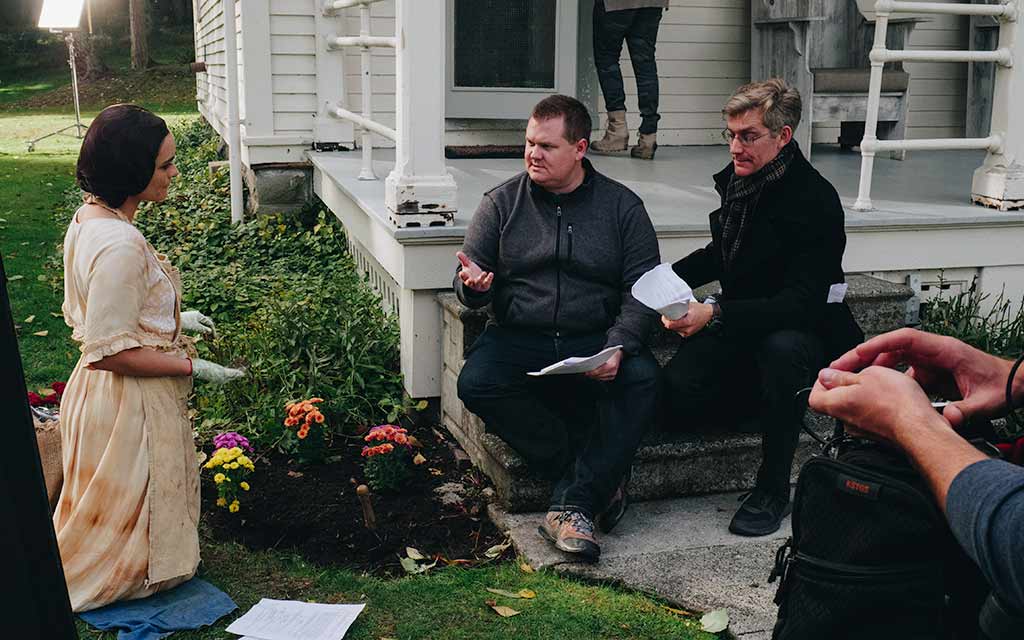 An actress is kneeling in a garden and two directors are instructing her how to play the scene.