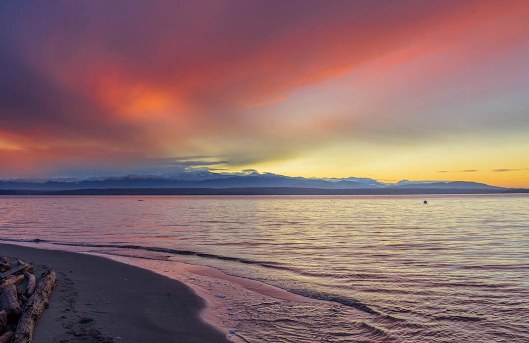 A beach and water with the setting sun and mountains in the distance and colorful clouds overhead.