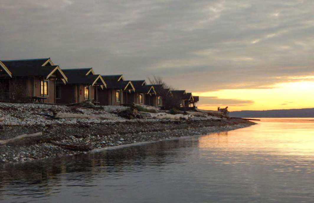 A row of one room cabins faces the water at sunset.