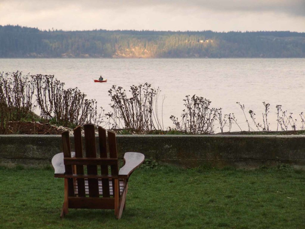 An empty wooden chair on a lawn looks out at a boat in the water.