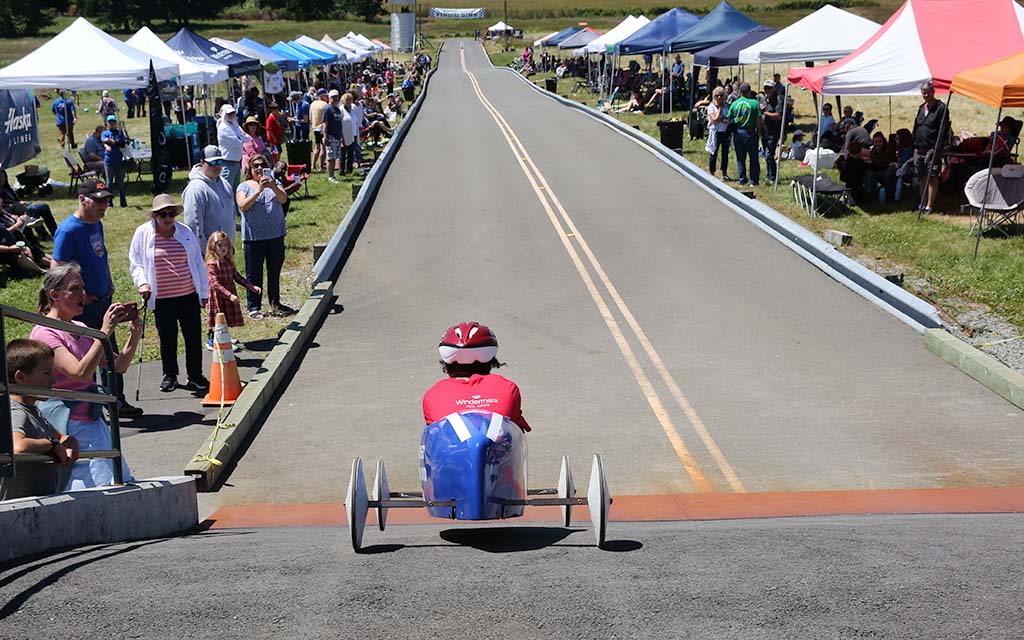 Soap Box race car at the starting line of a straignt and downhill race track with people watching on either side.