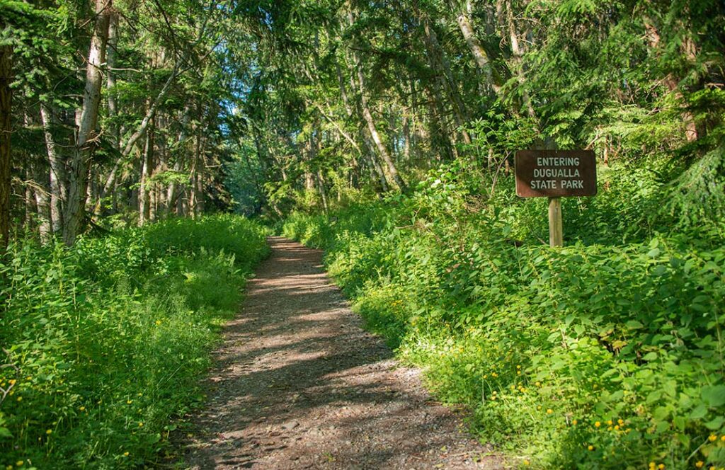 A wide trail into the woods with a small wooden sign reading "Dugualla State Park."