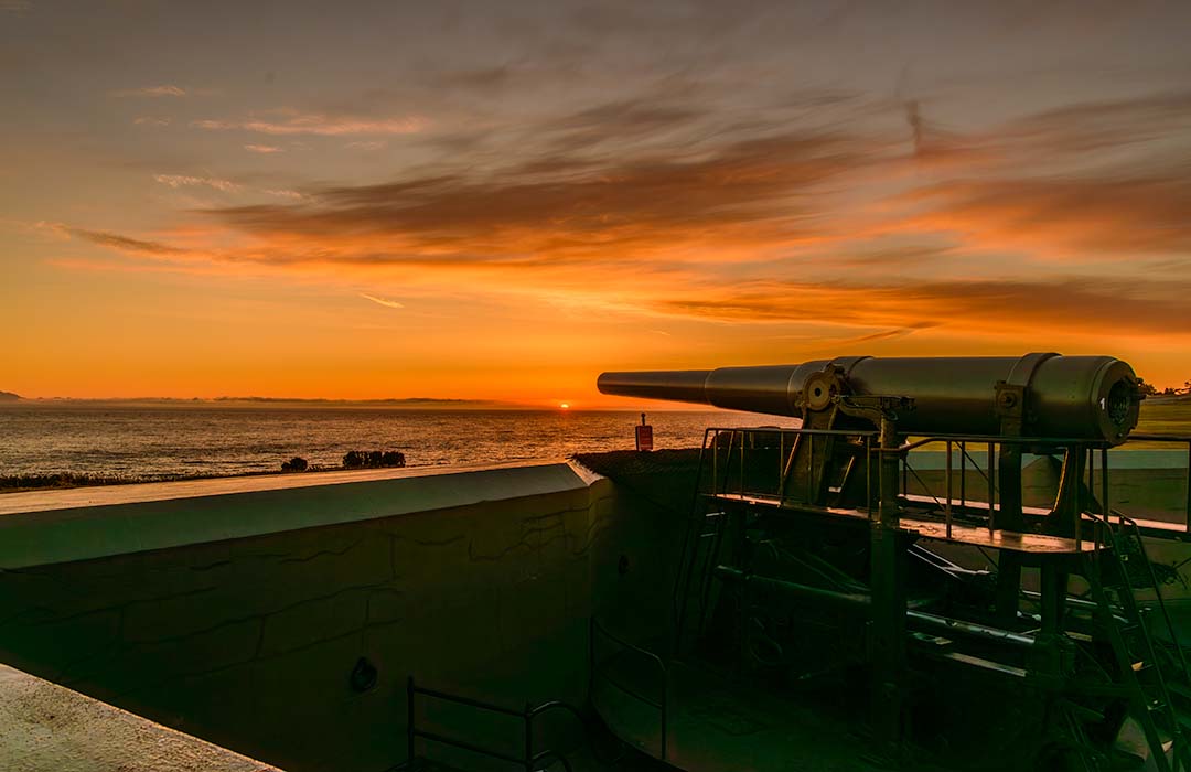 A large our-of-service cannon pointed out to the ocean with the sun setting behind it.