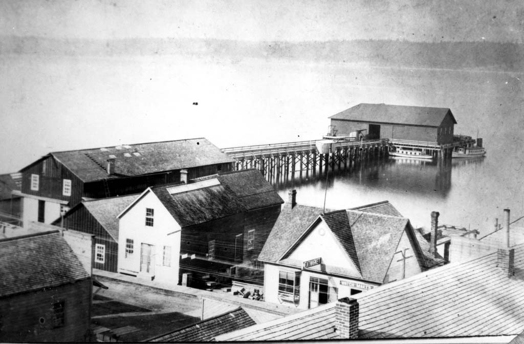 This is an old photo that is an aerial view of several wooden buildings and a wharf extending into the water.