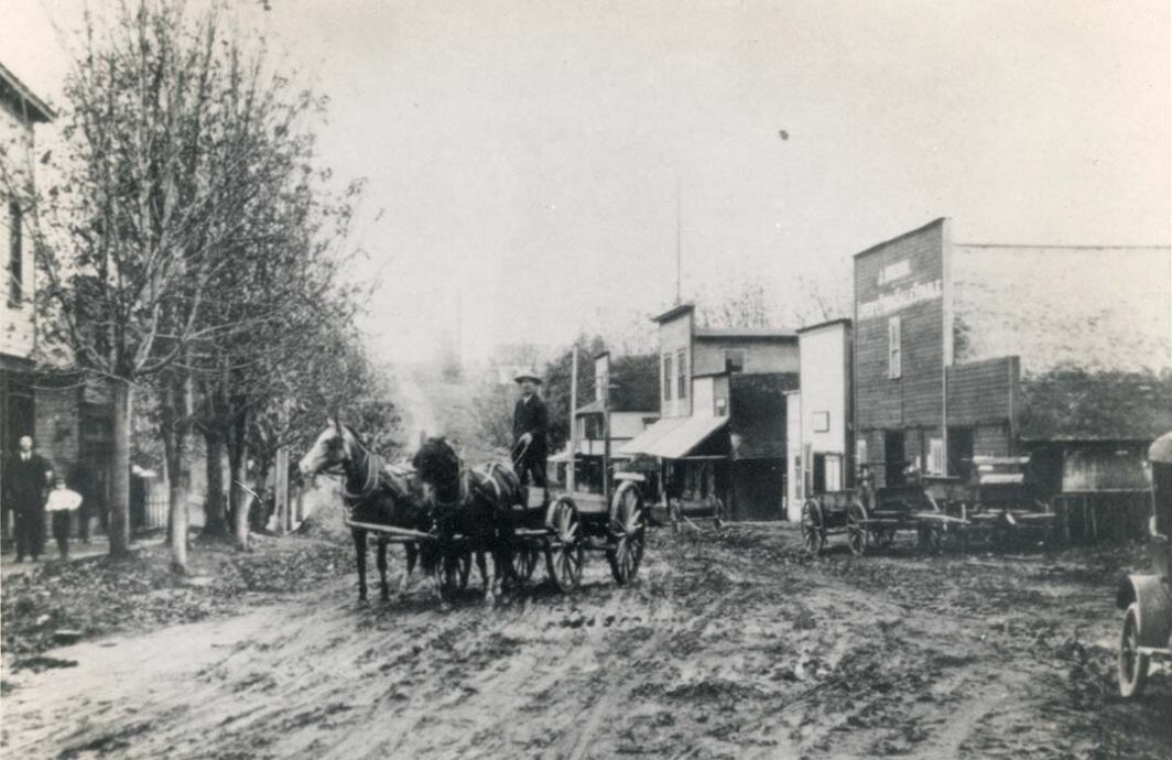 Old photo of a man standing in a wagon pulled by horses and they are on a muddy street lined with wooden buildings.