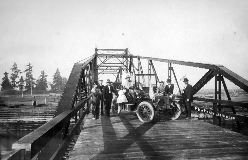 Old photo of a car and people on a small bridge.