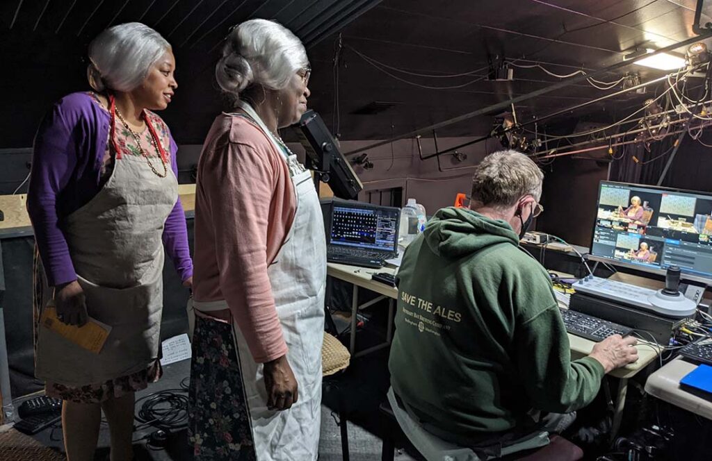 Two women in old-lady costumes watch a man edit a video of them on a computer.