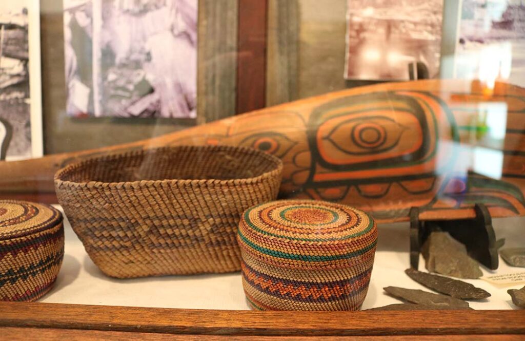 Baskets, arrowheads, a painted canoe paddle sit in a glass display case.
