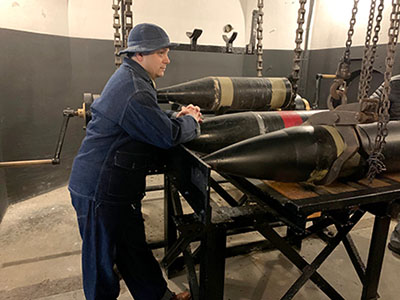 Man in an old-style uniform rests his hands on a large military shell.