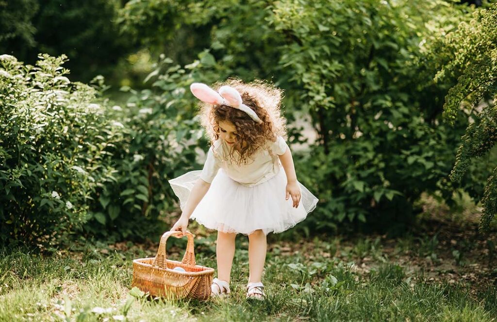 Young girl in a white dress and wearing rabbit ears is picking up a basket filled with eggs.