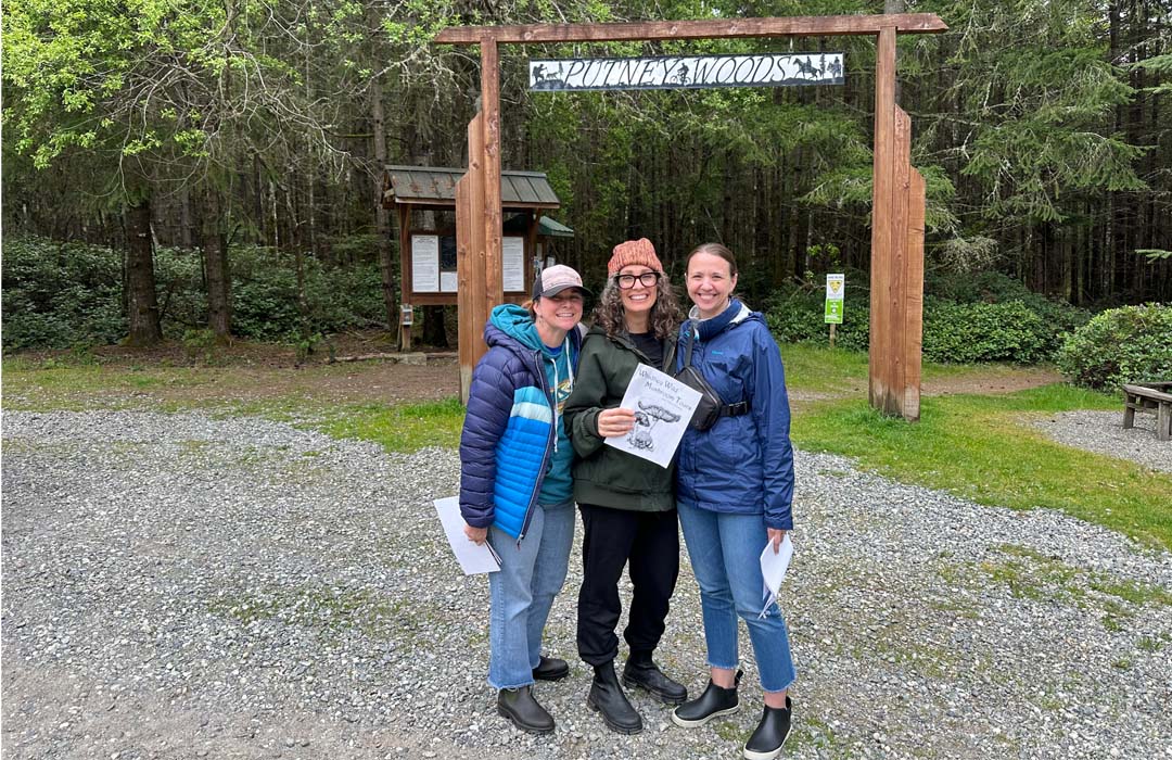 Three women posing for a picture in front of the entrance to Putney Woods.