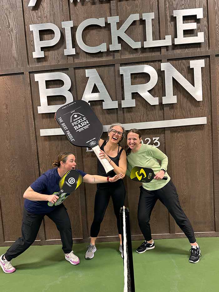 Three women mugging for the camera holding various sized pickle ball paddles.